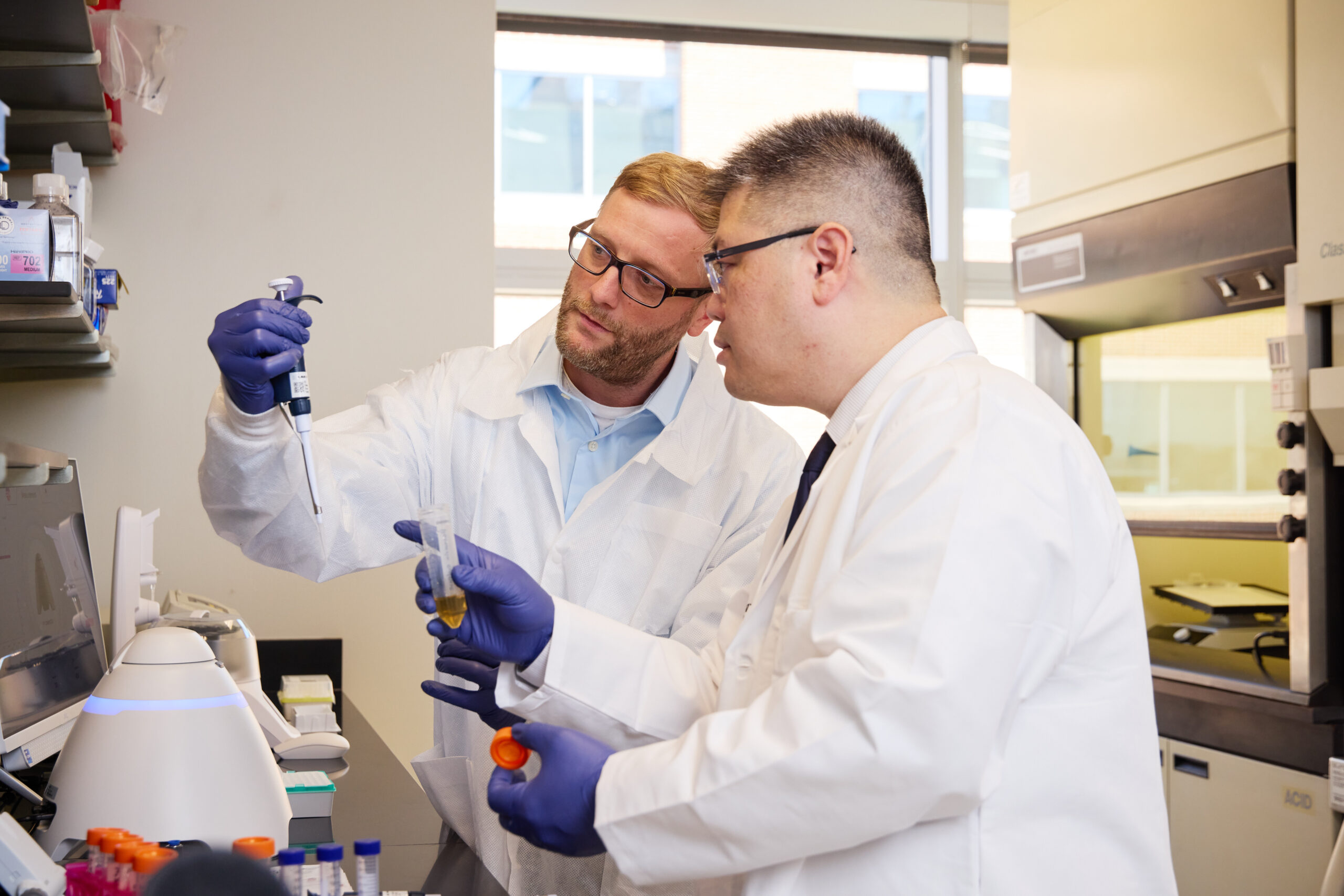 Two males in white coats and purple gloves in a lab.