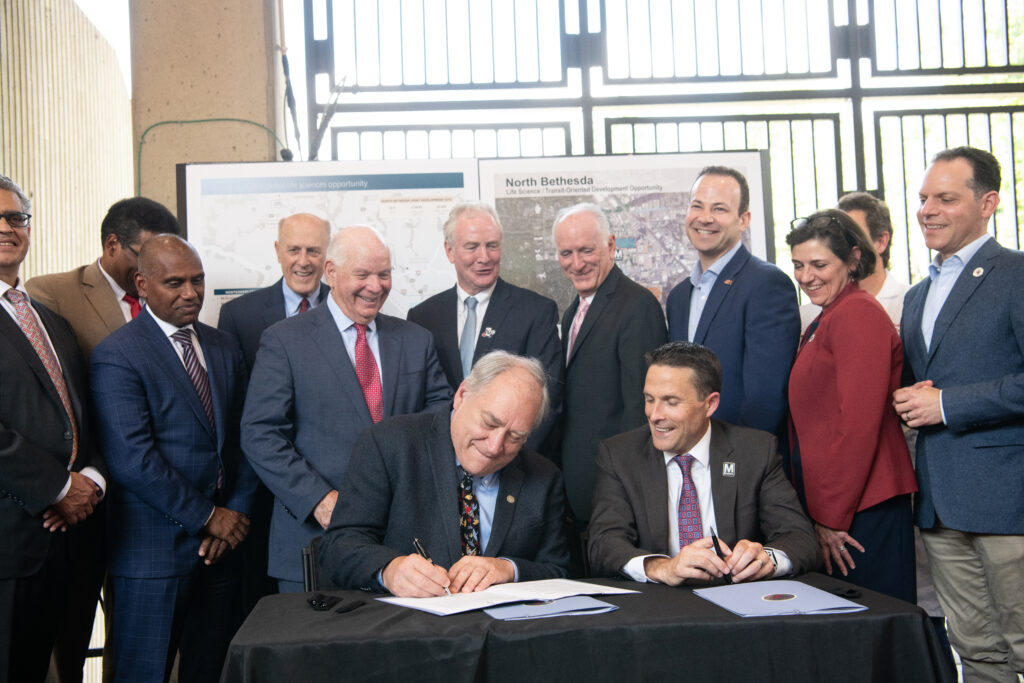 County executives gathered together. Two males seated in front, signing paperwork.