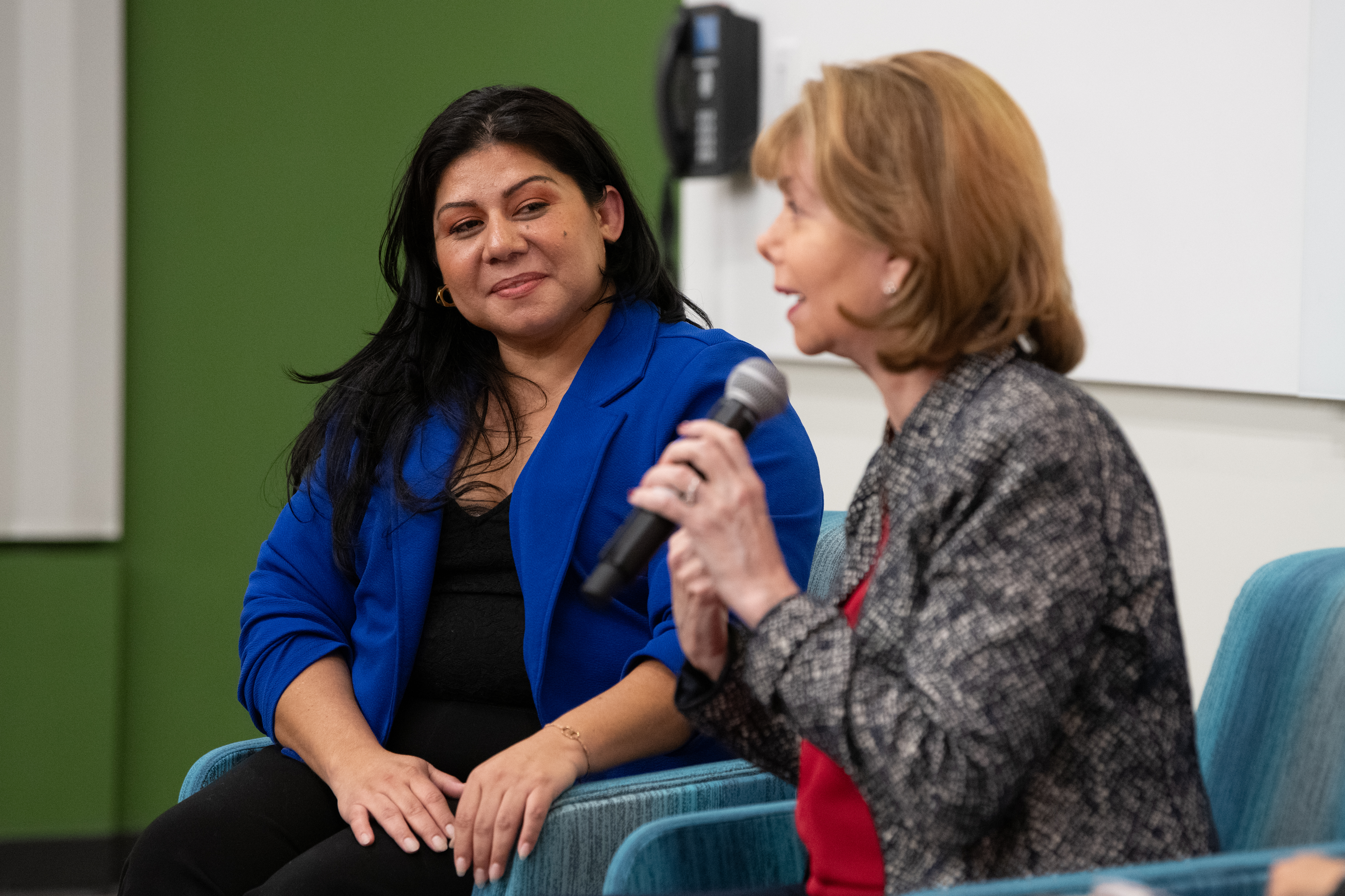 Women speaking with microphone as woman seated by her looks on.