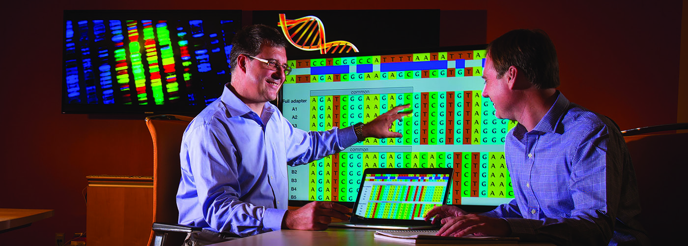 Two people analyzing colorful genetic data on screen and laptop.