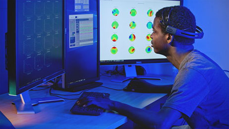 Blue-tinted photo of an African American male seated in front of two computer screens.
