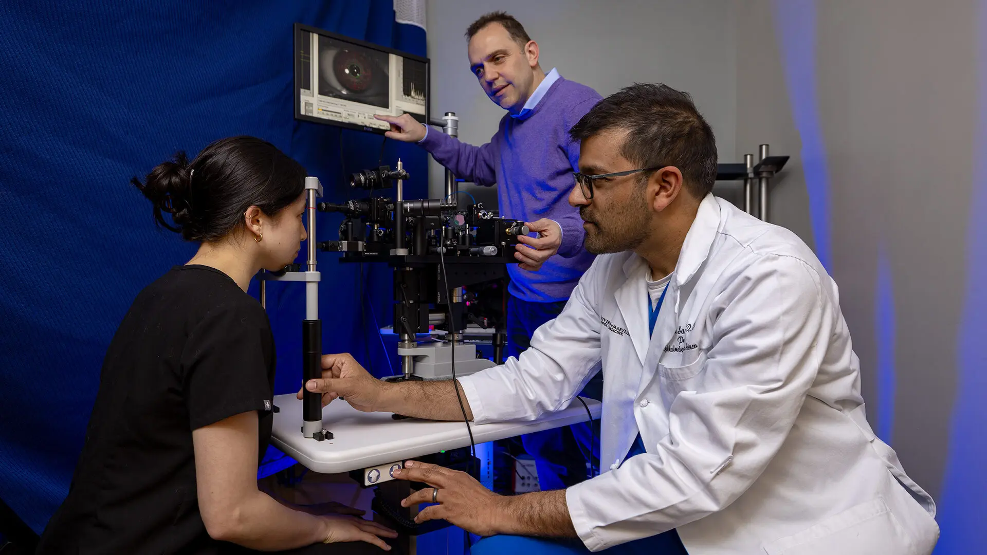 Healthcare professional in a white lab coat conducts an eye examination, while another person operates a computer monitor in the background.