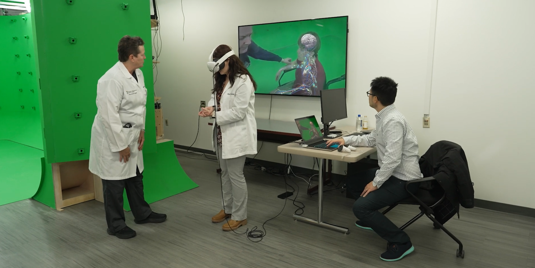 Three individuals in white lab coats pictured in front of a green screen and TV. One person pictured with virtual reality eye goggles.